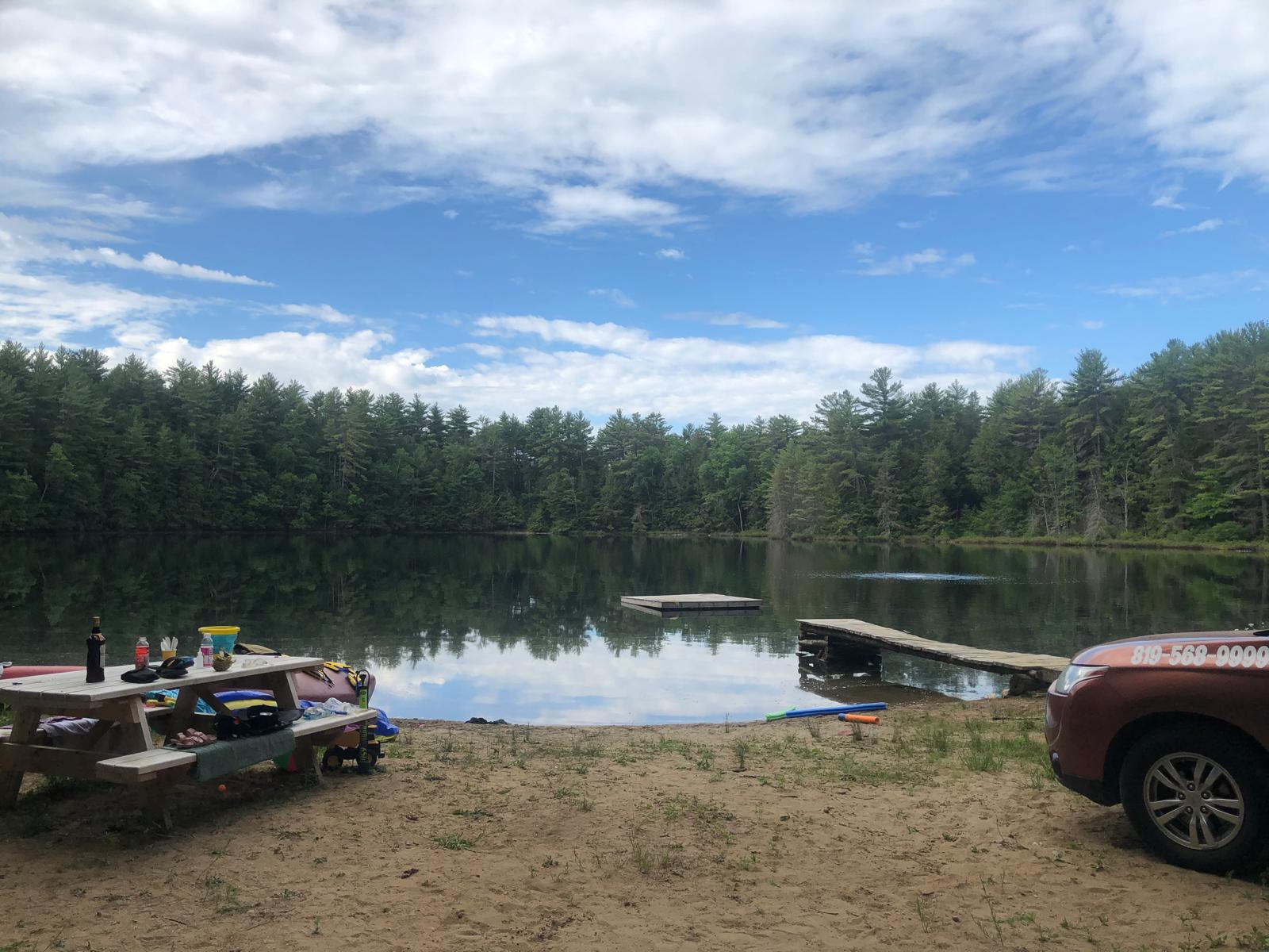 Sandy lakeside clearing with picnic tables and docks beneath a blue sky