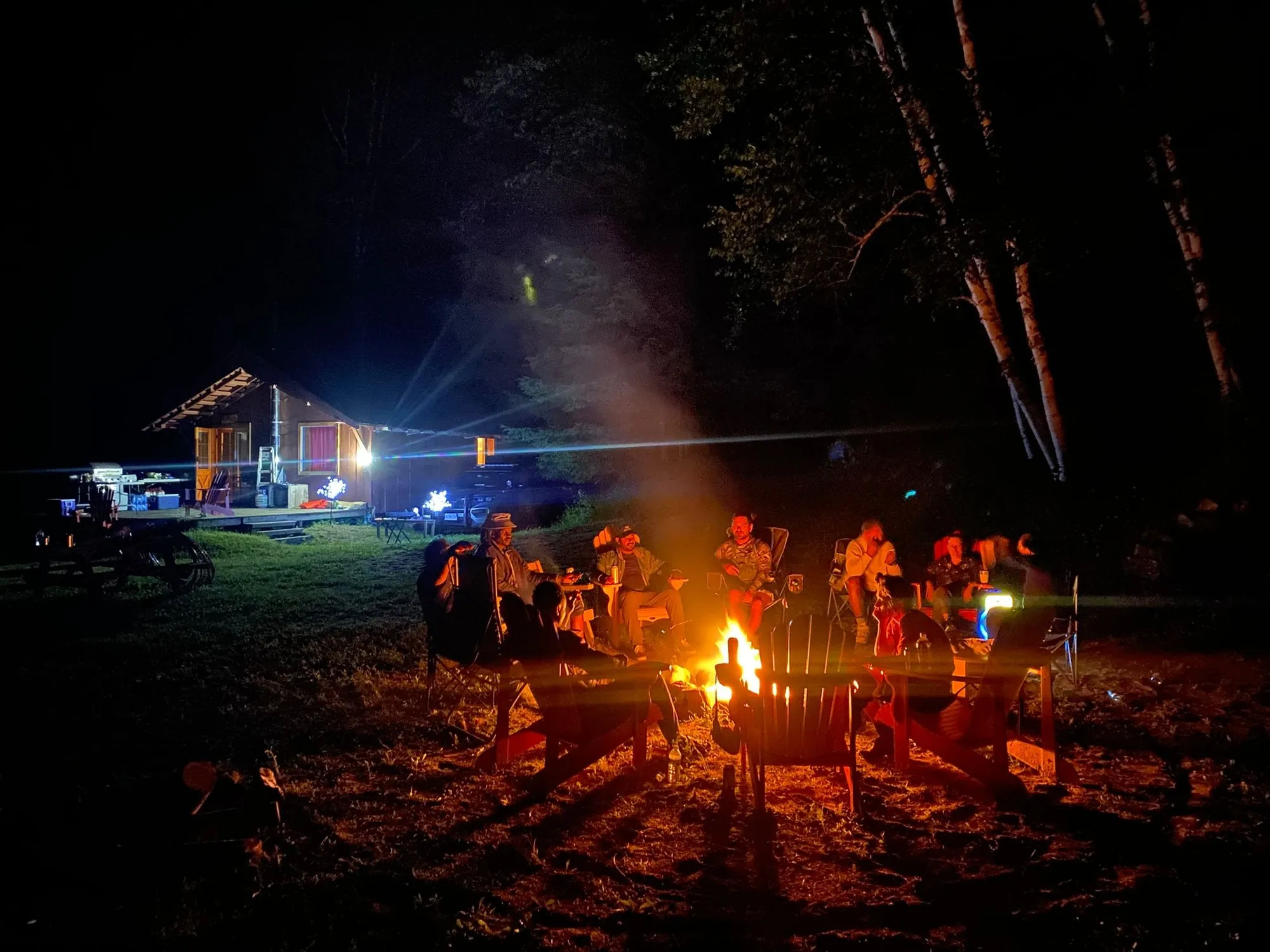 Guests gathered around a glowing campfire at night beside a lit cabin