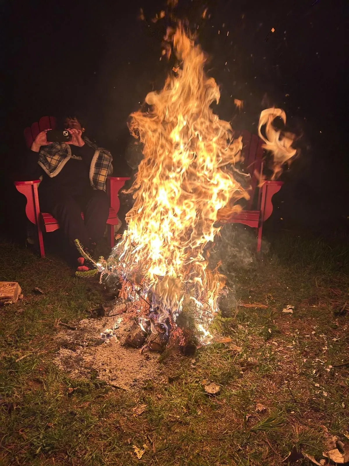 A tall bonfire blazing at night with red chairs around it