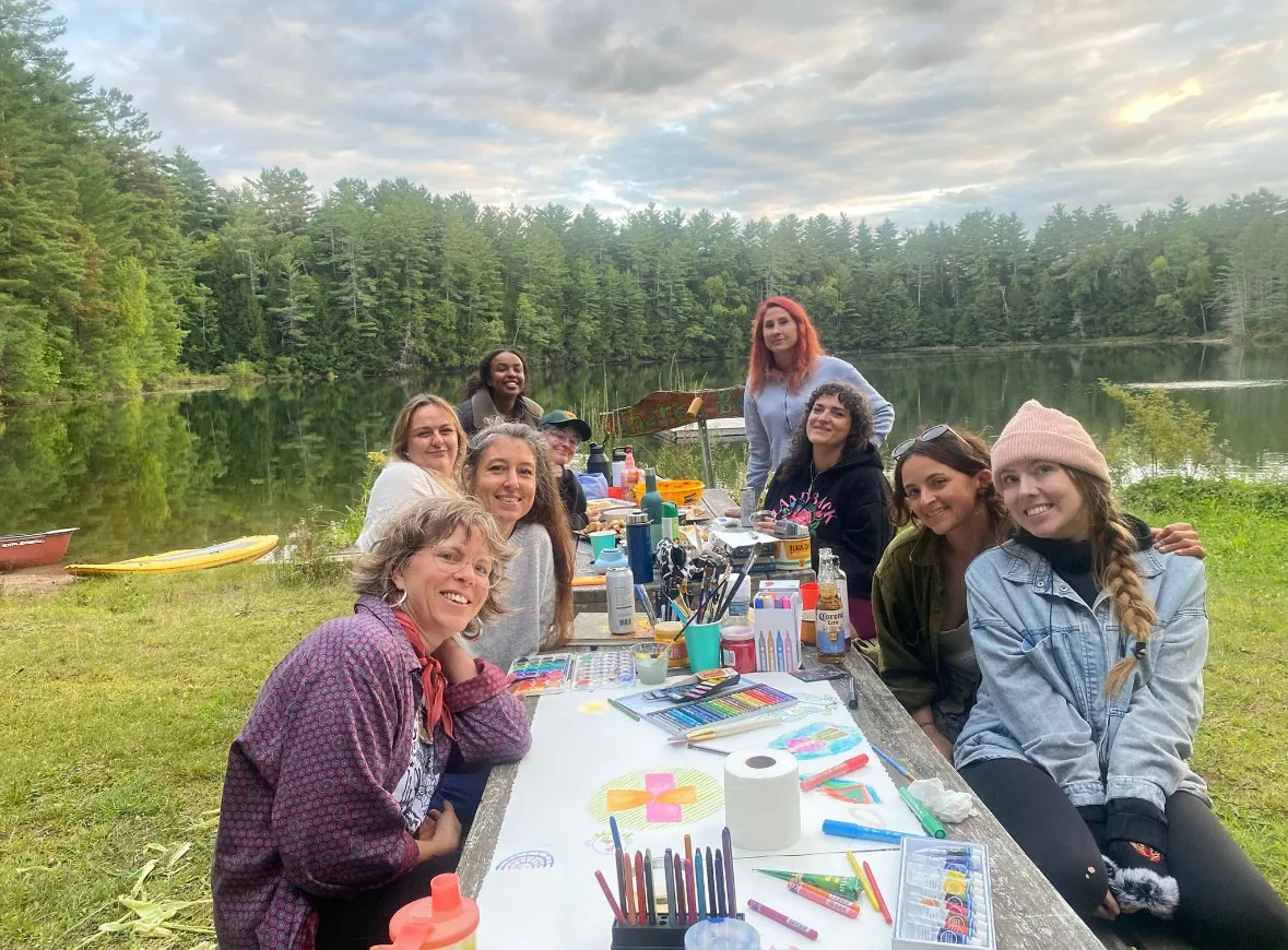 Friends gathered around a long lakeside table covered in art supplies