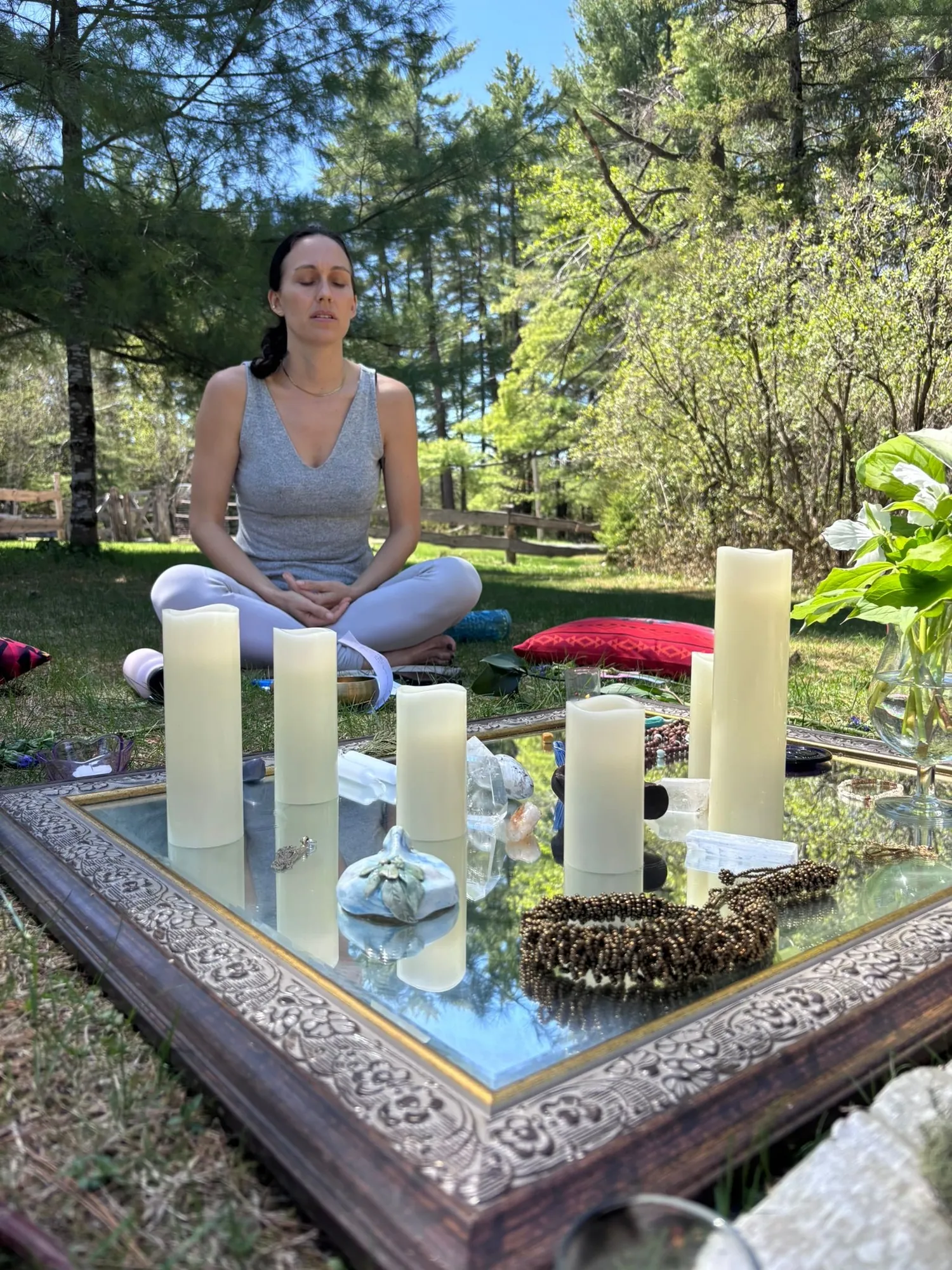 A woman meditating in the meadow behind a mirror altar of pillar candles and flowers